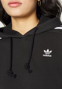 Svart hoodie med snörning och broderad vit Adidas-logotyp. Har kontrasterande vita ränder på axlarna och mjuk textur.
