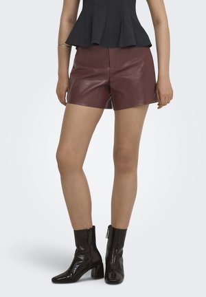 Vrouw draagt bruine leren shorts, zwarte peplum top en zwarte textuur enkellaarsjes, staat met ontspannen handen langs haar zij.