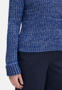 Pull en tricot dans des teintes variées de bleu, présentant une surface texturée et des poignets côtelés. Le design est ample avec un ourlet arrondi.