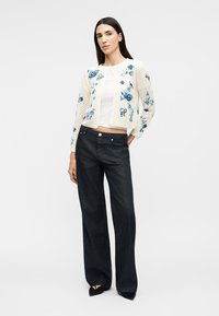 Kvinde iført en hvid, blomstermønstret strikket cardigan, hvid cropped top, sorte viddebenbukser og sorte spidse sko, stående med hånden i lommen.
