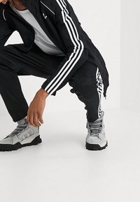 Černá sportovní bunda a kalhoty s bílými pruhy, šedé tričko a šedé boty se strukturovaným vzorem a černou podrážkou.