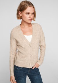 Beige gebreide cardigan met een V-hals, ribbelstructuur en drie grote knopen. Gepaarde met donkerblauwe jeans, wat een casual uitstraling geeft.