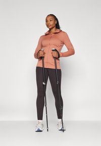 Mulher com um casaco rosa de fecho éclair, leggings pretas e ténis brancos, segurando dois paus de caminhada pretos contra um fundo claro.