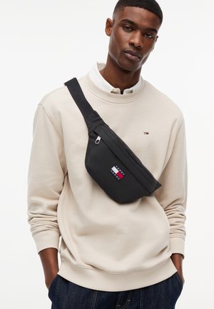 Mies, joka pitää päällään beigeä collegepaitaa ja tummat farkut, kantaa mustaa Tommy Jeans -olkalaukkua, jossa on vetoketjullinen etutasku.