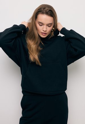 Kobieta o długich jasnobrązowych włosach poprawiająca kaptur swojej czarnej, oversize'owej bluzy na gładkim jasnym tle.