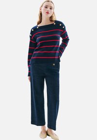 Pull rayé bleu marine avec des lignes rouges, ornée de boutons dorés ronds sur l'épaule ; associé à un pantalon en denim noir ample et des ballerines de couleur claire.