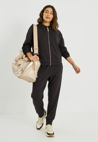 Sweat à capuche zippé noir et joggings amples, associés à un sac beige léger. Le mannequin porte des baskets crème et noir. Texture de tissu lisse.