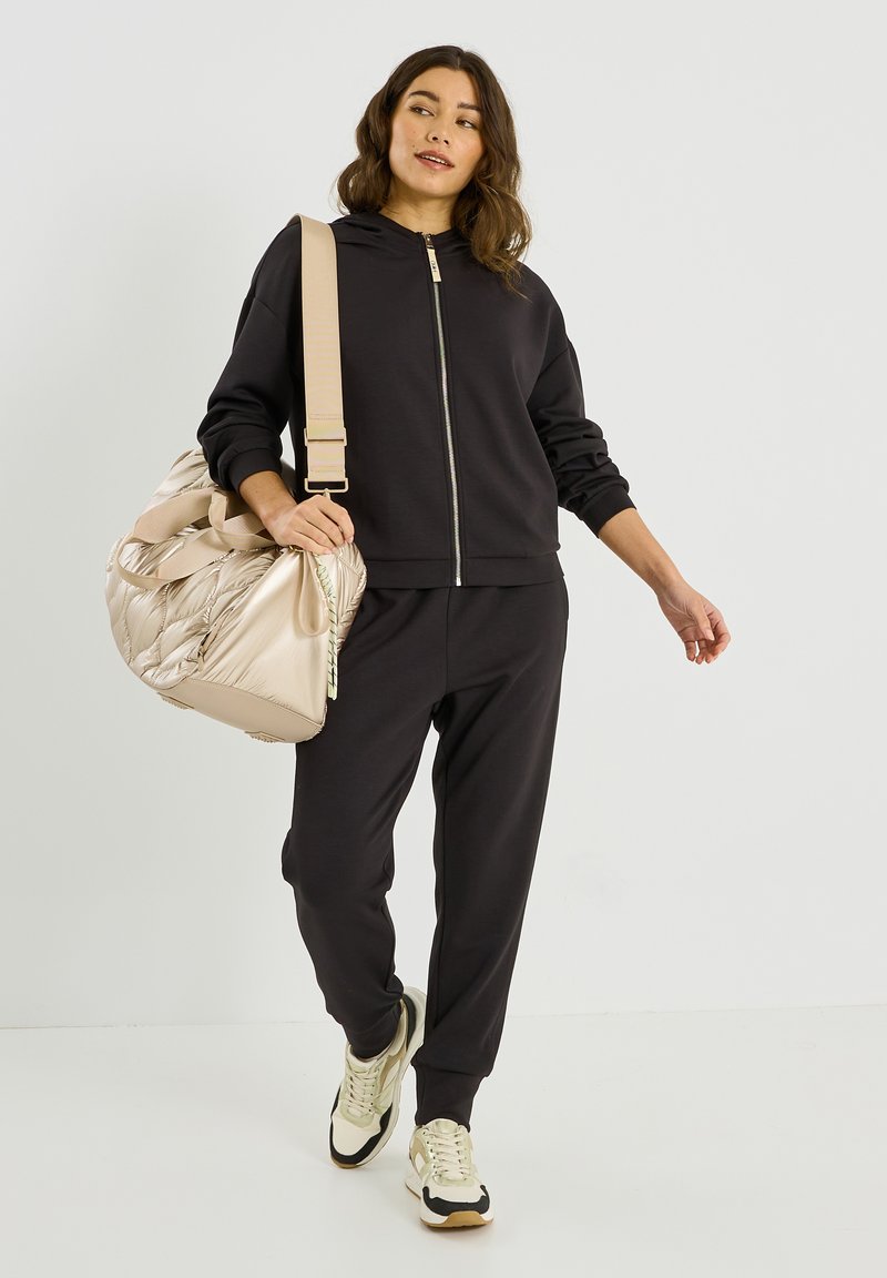 Sweat à capuche zippé noir et joggings amples, associés à un sac beige léger. Le mannequin porte des baskets crème et noir. Texture de tissu lisse.