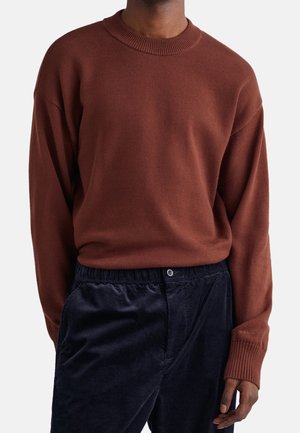 Homme portant un pull en laine marron à manches longues et un pantalon en velours côtelé bleu foncé, debout avec une main détendue le long du corps.