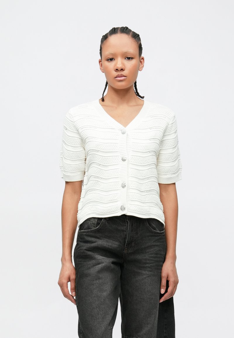 Γυναίκα με πλεκτά μαλλιά που φοράει λευκό υφασμάτινο κοντομάνικο ζακέτα με κουμπιά και άνετο σκούρο γκρι τζιν, στέκεται μπροστά σε απλό φόντο.