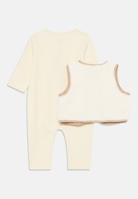 Une combinaison côtelée de couleur crème avec des manches longues et un col rond, associée à un gilet en fausse fourrure blanche. Le gilet a des accents de bordure beige.