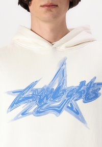 Sudadera blanca con un gran gráfico azul en forma de estrella y texto estilizado en el centro, hecha de una tela suave con una textura lisa.