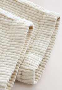 Tissu crème texturé avec des rainures verticales, doux au toucher, présentant un bord plié pour un effet superposé.