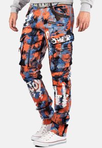 Pantalons cargo multicolores en orange, bleu et blanc avec des poches zippées, un tissu texturé et des accents graphiques. Portés avec des baskets blanches.