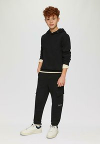 Schwarzer Kapuzenpullover mit Rundhalsausschnitt und hellbeigen Akzenten an den Bündchen, kombiniert mit schwarzen Cargo-Sweatpants und weißen Sneakers mit marineblauen Akzenten.
