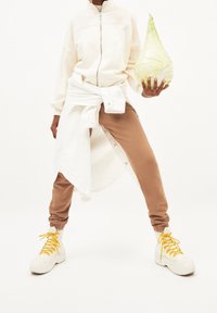 Felpa bianca con zip, pantaloni jogging beige e sneakers bianche chunky con lacci gialli. In mano una grande verza verde. Sfondo bianco.