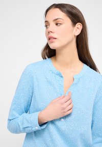 Hellblaues Hemd mit einem subtilen weißen Punktmuster, rundem Halsausschnitt und langen Ärmeln. Verfügt über Knopfdetails am Halsausschnitt und an den Manschetten.