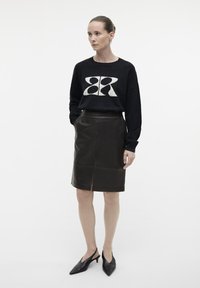 Sort strik Sweater med creme grafisk detalje, parret med en knælang mørkebrun lædernederdel med en midtsøm og forkantsslids. Sorte spidse hæle.