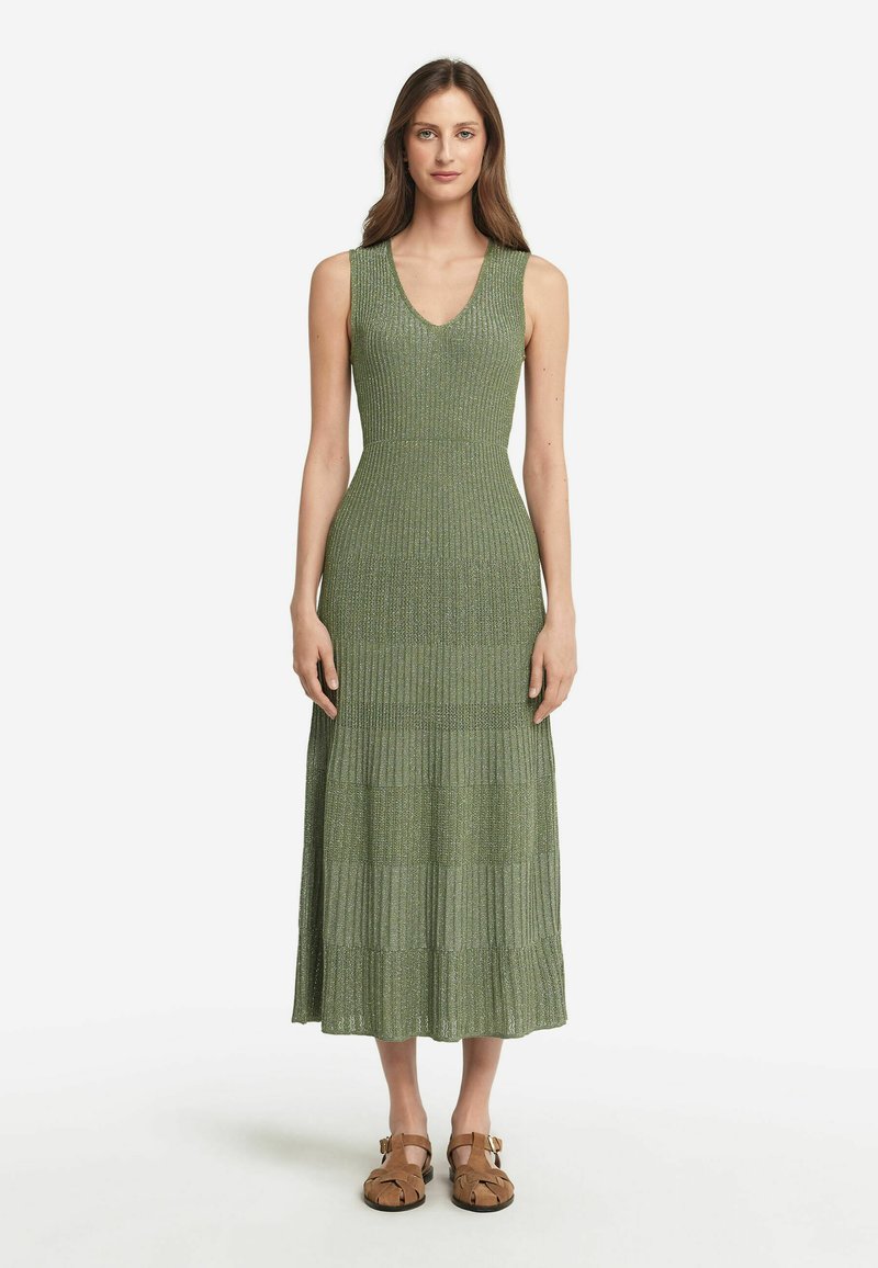 Robe verte sans manches, arrivant aux genoux, avec un effet côtelé et un ourlet en tiers. Portée avec des sandales marron à brides réglables.