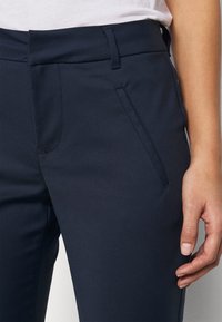 Pantaloni in tessuto elasticizzato blu navy con vita piatta, tasche laterali e vestibilità sartoriale. Texture liscia con dettagli di cucitura minimi.
