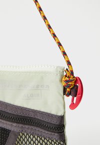 Funda de malla verde y gris con cremallera superior, base reforzada y correa anudada multicolor. Presenta un clip rojo para su sujeción.