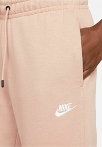 Ljust rosa Nike sweatpants med dragsko i midjan, sidfickor och vitt broderat logotyp på den nedre benet. Mjuk tygtextur.
