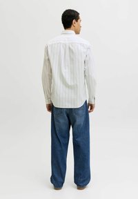 Homme debout de dos, portant une chemise blanche à rayures à manches longues et un jean bleu ample, sur un fond clair uni.