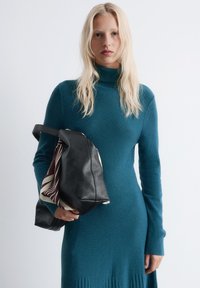 Junge Frau mit langen blonden Haaren, die ein türkisfarbenes Turtleneck-Kleid trägt, hält eine schwarze Lederhandtasche mit einem gestreiften Schal darin.