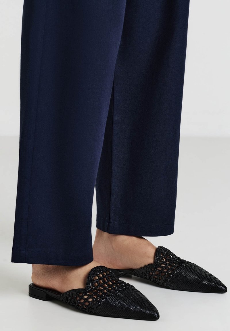 Des mules noires à bout pointu présentent une texture tissée et des accents lisses. Associées à un pantalon ample bleu marine qui couvre les chevilles.