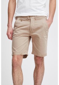 Homme portant un short beige jusqu'aux genoux avec une fermeture à bouton et à zip, poches avant, et un t-shirt blanc, se tenant avec une main dans la poche.