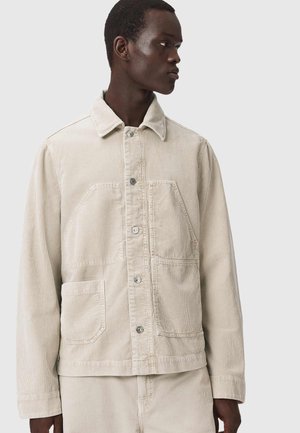 Mand iført beige kordjakke og matchende bukser, kigger til venstre mod en ensartet lysegrå baggrund.