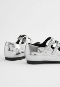 Silberne metallische Schuhe mit einer glatten Oberfläche, ausgestattet mit zwei Schnallenriemen, runden Spitzen und einer flachen schwarzen Sohle.