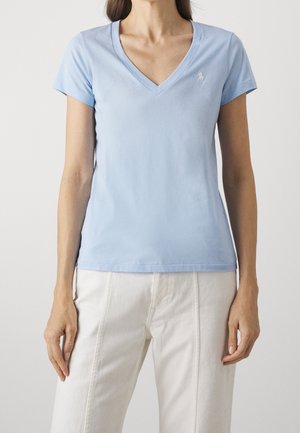Mujer con camiseta azul claro de manga corta y escote en V y pantalones color beige claro, de pie con los brazos relajados a los lados, sobre un fondo liso.