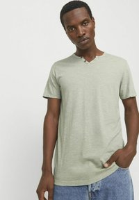 Lys grøn kortærmet T-shirt med V-udskæring og detaljer med to knapper, lavet af et struktureret stof. Bæret med blå denim shorts.