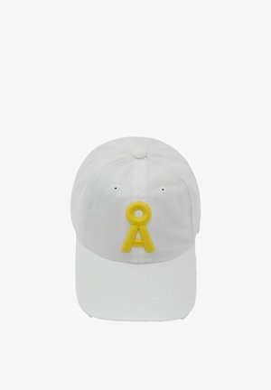 Donna con lunghi capelli scuri indossa un berretto bianco con la lettera A gialla, una maglietta bianca a maniche corte a coste e piccoli orecchini a cerchio dorati, espressione neutra.
