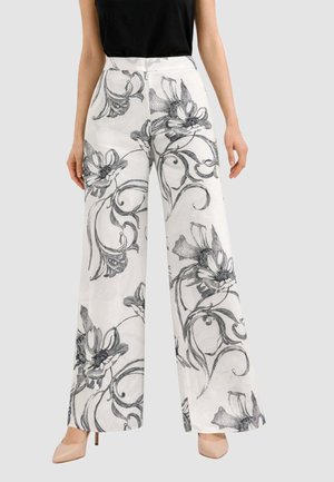 Donna che indossa pantaloni bianchi a vita alta e gamba larga con grandi motivi floreali neri e vortici, abbinati a tacchi color nudo e un top nero senza maniche.