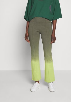 Person iført grønne ombré vidde bukser, hvide sneakers og en løs mørkegrøn top, stående foran en ensfarvet lys baggrund.