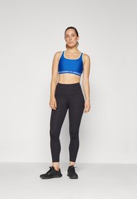 Blue sports bra with logo band, paired with black leggings. Smooth texture, form-fitting design, suitable for athletic activities, with black sneakers.