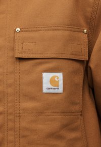 Καφέ τζάκετ από ανθεκτικό ύφασμα με τσέπη και καπάκι ασφαλισμένο με μεταλλικές ξυλόβιδες και λευκή ετικέτα με το λογότυπο Carhartt στο κέντρο.