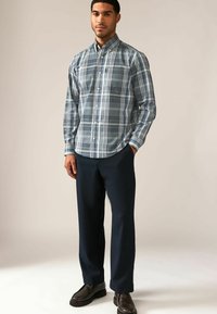 Geruit button-up shirt in tinten van blauw en grijs; lange mouwen, kraag en een afgeronde zoom. Gecombineerd met donkere broeken en zwarte schoenen.