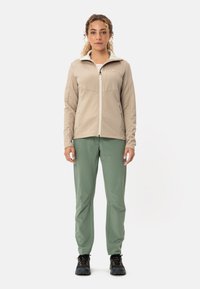 Giacca leggera beige con zip e motivo testurizzato, abbinata a pantaloni verdi a sigaretta e scarpe da trekking nere, in piedi su uno sfondo bianco.