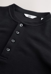 Chemise en tissu côtelé noir à col rond avec quatre boutons foncés le long du plastron avant, affichant l'étiquette intérieure indiquant "NEXT EDIT."