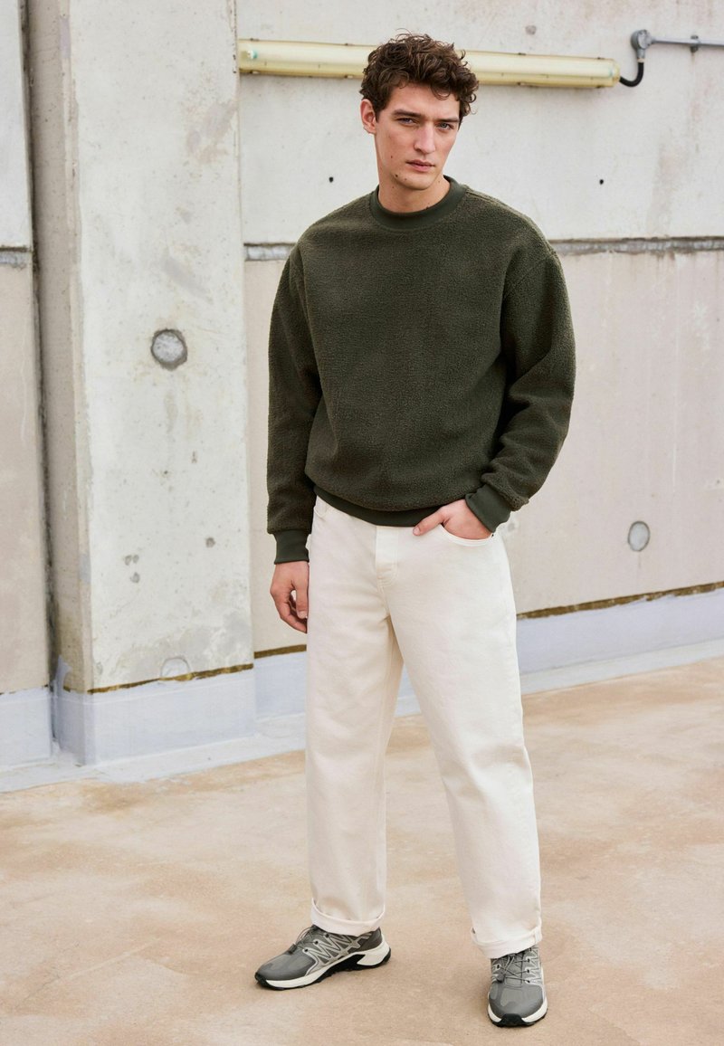 Olivgrüner Fleece-Pullover, weiße Jeanshose und graue Turnschuhe. Das Outfit hat eine lockere Passform und gerippte Akzente an den Bündchen und am Saum.