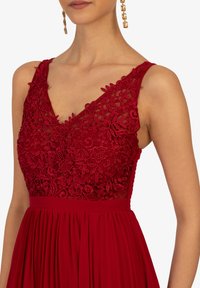 Abito rosso con scollatura a V, caratterizzato da dettagli in pizzo floreale sul corpetto e una gonna plissettata. Accentuato da una vita aderente.