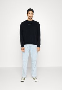 Felpa nera con logo bianco, colletto a giro, e polsini a costine. Abbinata a jeans azzurri chiari e scarpe da ginnastica verdi con suola bianca.