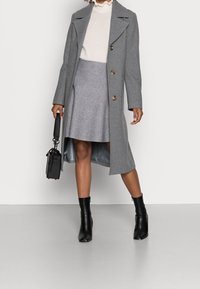 Abrigo de lana gris con botones grandes, combinado con una falda gris clara. Botas negras de tacón alto y un pequeño bolso negro completan el conjunto.