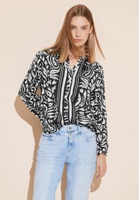 Blusa a maniche lunghe in bianco e nero con motivi floreali e a righe, abbinata a jeans di denim blu chiaro. Tessuto morbido e dettagli con bottoni visibili.