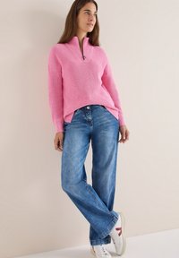 Maglione rosa lavorato a maglia con zip e maniche ampie, abbinato a jeans a gamba larga blu e sneakers bianche con accenti rossi. Sfondo semplice.
