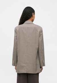 Femme aux longs cheveux bouclés portant un blazer oversize en pied-de-poule marron et un pantalon marron foncé, de dos contre un fond uni.