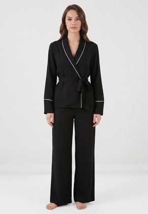 Kvinde stående barfodet iført sort wrap-style pyjamasæt med hvide kanter på krave og manchetter mod ensfarvet baggrund.